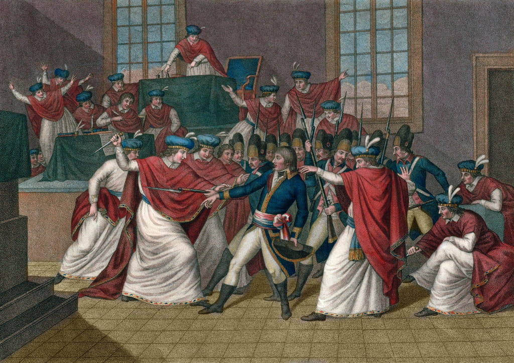 This colored engraving captures a pivotal moment during the French Revolution, specifically the Coup d’État of 18 Brumaire by Napoleon Bonaparte. The scene is set in an austere room with high windows, through which daylight pours. It depicts a chaotic confrontation between various groups. In the foreground, two men in white robes with red drapery, representing members of the government, are being arrested or coerced by military officers in blue uniforms with white trousers and bicorne hats. One government member clutches a sheathed sword, symbolizing his power being overtaken. Behind them, other officers and government officials are engaged in a heated dispute, some with swords drawn. To the left, a group of men in red judicial robes gesture dramatically, indicating a scene of intense political upheaval. The overall composition conveys the tension and disorder of this historical event.