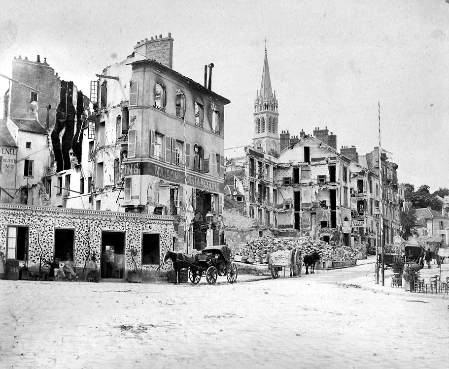 This historical black and white photograph depicts the aftermath of the bombing of Paris during the Franco-Prussian War. The scene shows a street with damaged buildings; some are partially collapsed, with visible rubble and destruction. The facades of the remaining structures are riddled with holes and show signs of bombardment. In the foreground, there's a horse-drawn carriage and some people going about their daily activities, indicating life continues amidst the ruins. A pile of debris is stacked by the side of the street, and in the background, an intact church bell tower rises above the devastation, contrasting with the surrounding destruction. The image captures a moment of resilience in a war-torn city.